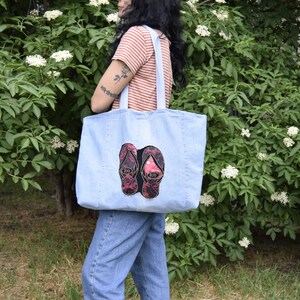 May include: A light blue denim tote bag with a pair of pink and black flip flops embroidered on the front.