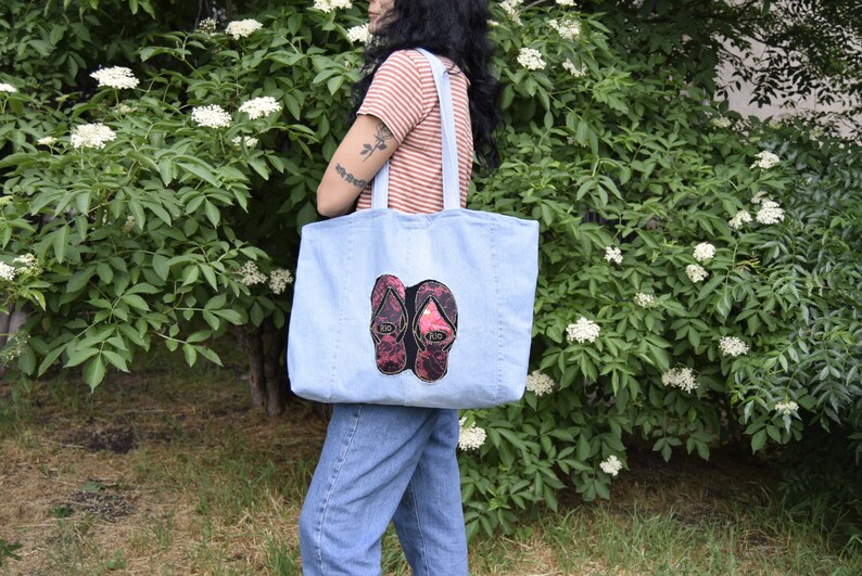 May include: A light blue denim tote bag with a pair of pink and black flip flops embroidered on the front.