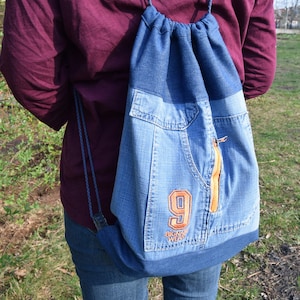 Op de afbeelding: Een blauwe denim trekkoordrugzak met een groot nummer "9" en de tekst "BIG NAME WEAR" geborduurd op de voorkant.