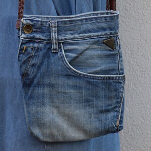 Puede incluir: Una peque&ntilde;a bolsa de mezclilla azul con una correa de cuero marr&oacute;n y una hebilla de metal. La bolsa tiene un bolsillo en la parte delantera.