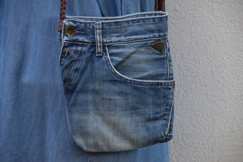 Puede incluir: Una peque&ntilde;a bolsa de mezclilla azul con una correa de cuero marr&oacute;n y una hebilla de metal. La bolsa tiene un bolsillo en la parte delantera.