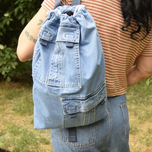 May include: A blue denim drawstring backpack with multiple pockets. The backpack is being worn by a person with long dark hair.