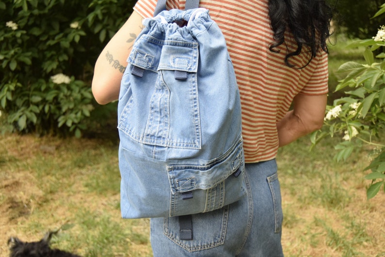 May include: A blue denim drawstring backpack with multiple pockets. The backpack is being worn by a person with long dark hair.