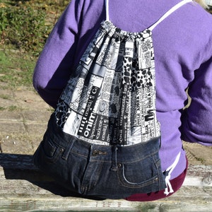 Könnte beinhalten: Eine schwarz-weiße Kordeltasche aus recyceltem Denim und Stoff mit Zeitungsdruck. Die Tasche hat eine weiße Kordel und wird von einer Person getragen.