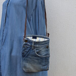 Puede incluir: Un bolso bandolera de mezclilla azul con una correa trenzada marr&oacute;n. El bolso est&aacute; hecho de mezclilla reutilizada y tiene un forro a rayas blanco y azul.