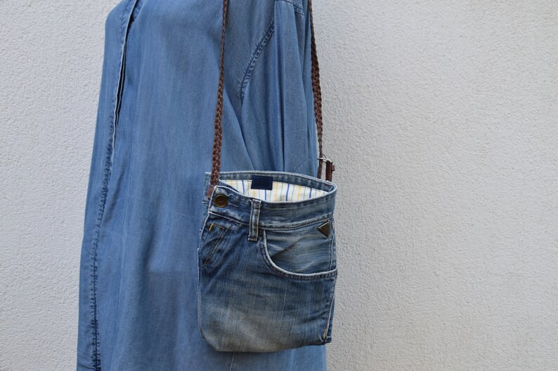 Puede incluir: Un bolso bandolera de mezclilla azul con una correa trenzada marr&oacute;n. El bolso est&aacute; hecho de mezclilla reutilizada y tiene un forro a rayas blanco y azul.