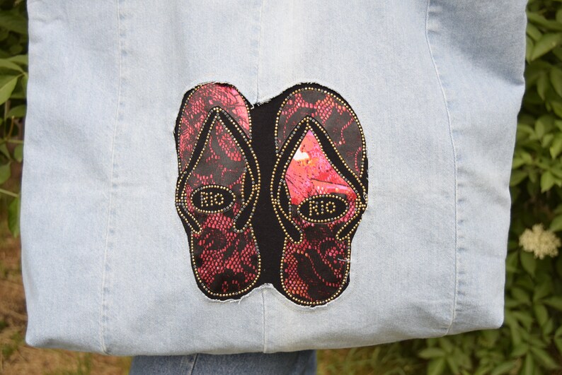 May include: A light blue denim tote bag with a black and red embroidered design of two flip flops. The flip flops have gold accents and the word "Rio" written on them.