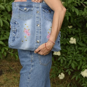 May include: A denim tote bag with embroidered pink flowers and a button closure. The bag is made from recycled denim and has a casual, vintage style.