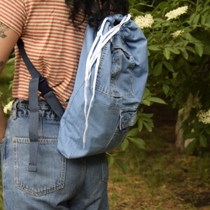 May include: A person wearing a striped shirt and blue jeans is carrying a blue denim drawstring backpack with white drawstrings.