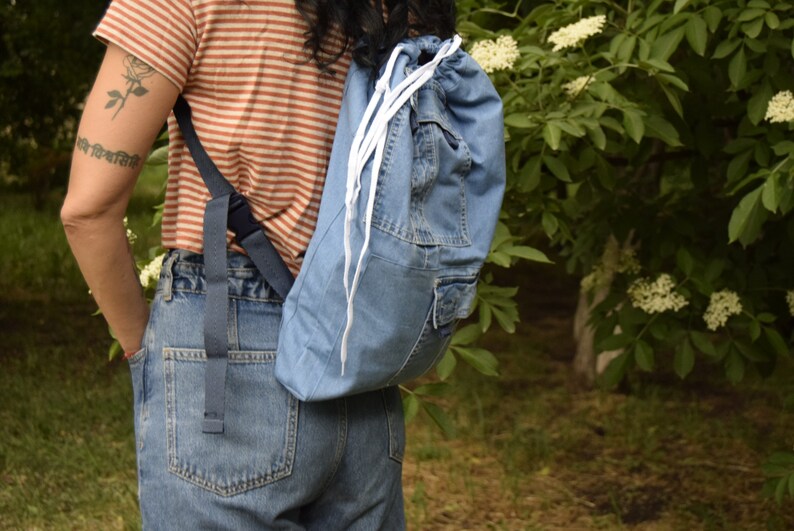 May include: A person wearing a striped shirt and blue jeans is carrying a blue denim drawstring backpack with white drawstrings.