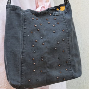 May include: A gray denim tote bag with a black and gold beaded design. The bag has a distressed look and is made from recycled materials.