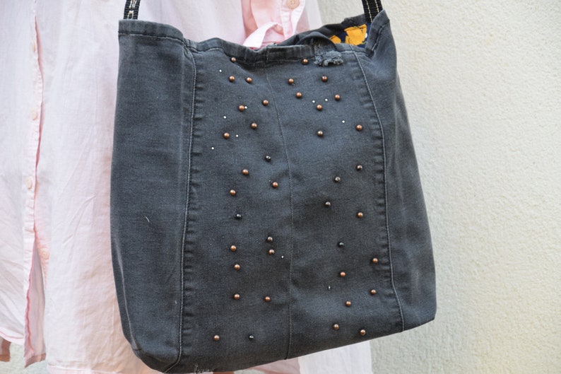 May include: A gray denim tote bag with a black and gold beaded design. The bag has a distressed look and is made from recycled materials.