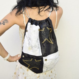 Può includere: Una borsa a cordoncino bianca e nera con dettagli a stella dorata. La borsa è realizzata con un patchwork di diversi tessuti e ha una stella nera con strass sul davanti.