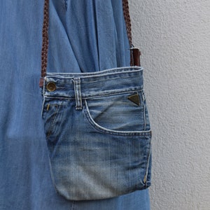 Puede incluir: Un bolso bandolera de mezclilla azul con una correa marr&oacute;n. El bolso est&aacute; hecho de mezclilla reutilizada y tiene un bolsillo frontal.
