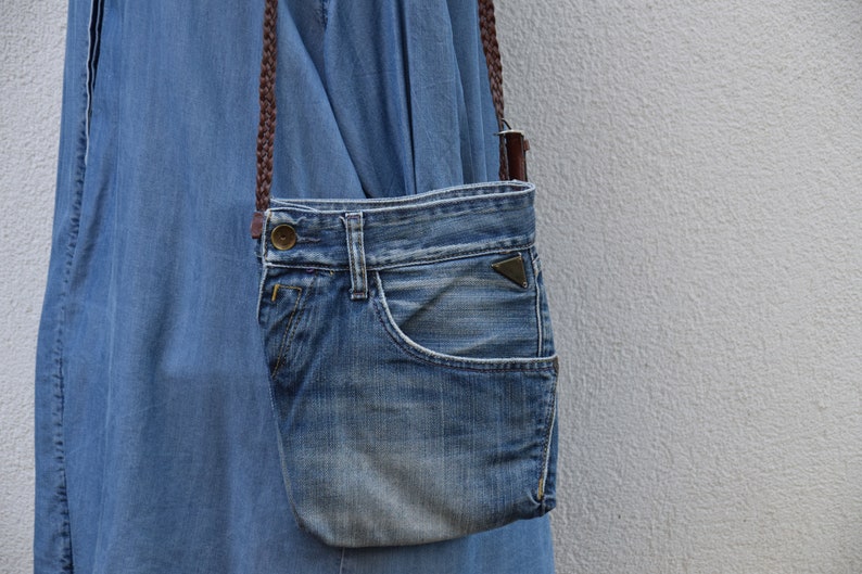 Puede incluir: Un bolso bandolera de mezclilla azul con una correa marr&oacute;n. El bolso est&aacute; hecho de mezclilla reutilizada y tiene un bolsillo frontal.