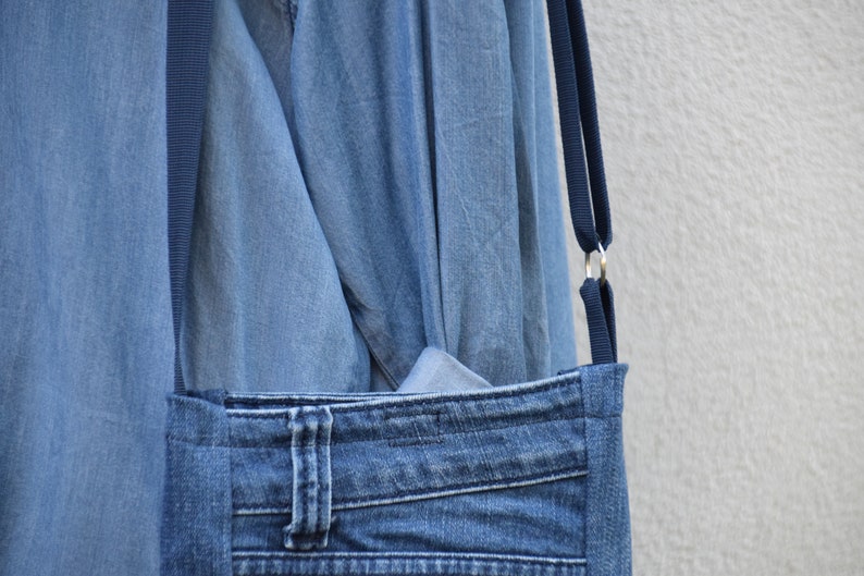 Puede incluir: Un bolso cruzado de mezclilla con una correa azul oscuro y anillos de metal plateado. El bolso presenta un bolsillo frontal hecho de jeans reciclados, incluidos los presillas y costuras originales. El bolso es de mezclilla azul claro.