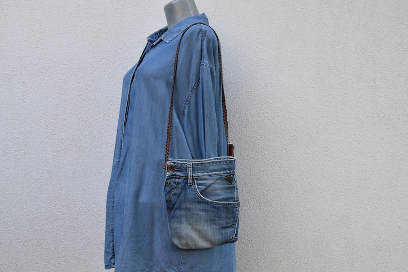 Puede incluir: Un bolso bandolera de mezclilla azul con una correa marr&oacute;n. El bolso est&aacute; hecho de mezclilla reutilizada y tiene un aspecto &uacute;nico y hecho a mano.