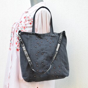 May include: A dark gray denim tote bag with a black and white strap that says "SHINECCI" on it. The bag is decorated with small, round, bronze-colored studs.