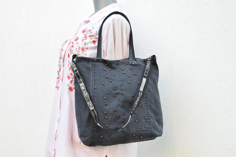 May include: A dark gray denim tote bag with a black and white strap that says "SHINECCI" on it. The bag is decorated with small, round, bronze-colored studs.