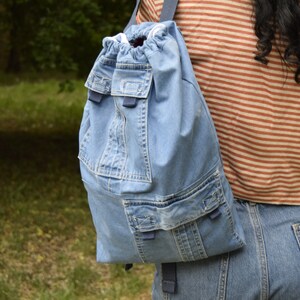 May include: A light blue denim backpack with a drawstring closure and multiple pockets. The backpack has a blue strap and is worn by a person.