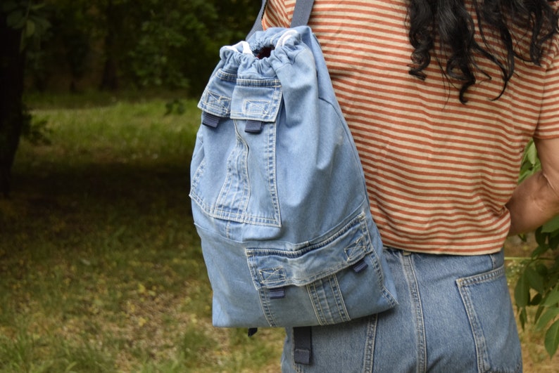 May include: A light blue denim backpack with a drawstring closure and multiple pockets. The backpack has a blue strap and is worn by a person.