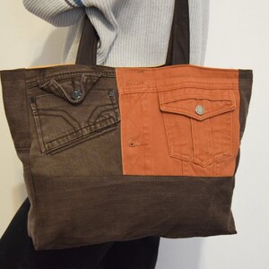 Może przedstawiać: Brązowa i pomarańczowa torba typu tote wykonana z odzyskanego denimu. Torba ma dwie duże kieszenie z przodu, jedną brązową i jedną pomarańczową.