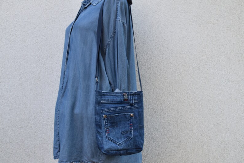 Puede incluir: Un bolso cruzado de mezclilla hecho de jeans reciclados. El bolso presenta un bolsillo trasero, trabillas para el cintur&oacute;n y un cierre de bot&oacute;n. El bolso tiene una correa larga y delgada y es de color azul medio.