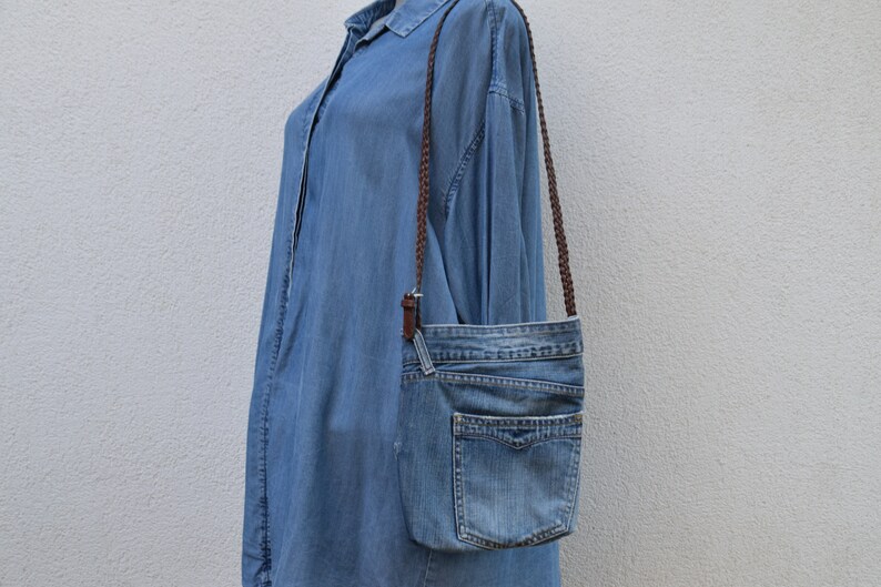 Puede incluir: Un bolso bandolera de mezclilla azul con una correa trenzada marr&oacute;n. El bolso tiene un cierre de solapa y un bolsillo grande en la parte delantera.