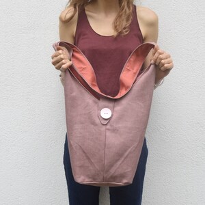 May include: A dusty rose colored tote bag with a large white button closure. The bag has a pink lining and is made of a soft, textured fabric.