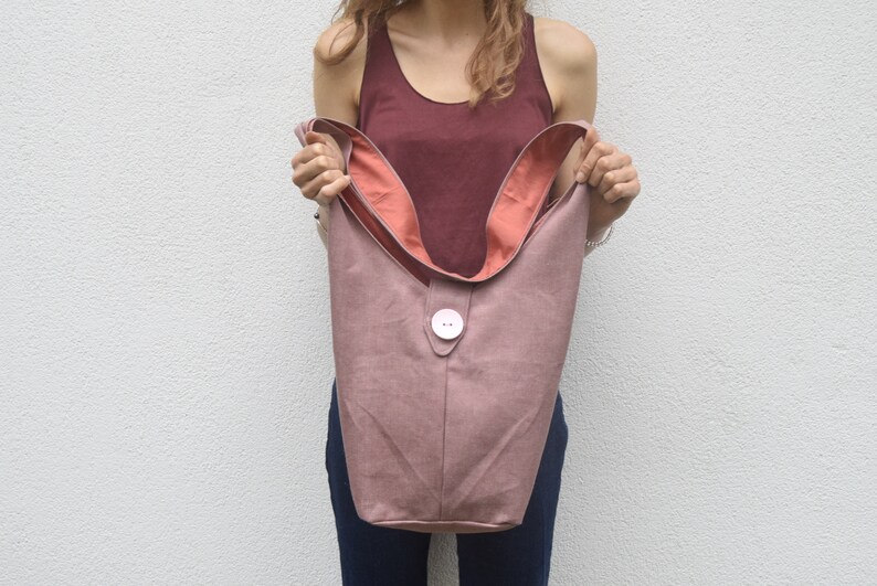 May include: A dusty rose colored tote bag with a large white button closure. The bag has a pink lining and is made of a soft, textured fabric.