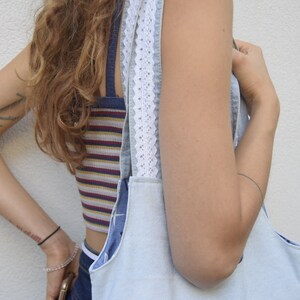 May include: A light blue denim tote bag with white lace trim on the straps. The bag is being held by a person wearing a striped tank top and jeans. The person has a tattoo on their arm that says "Home" with an anchor.
