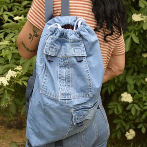 May include: A blue denim backpack with a drawstring closure and multiple pockets. The backpack is made from recycled denim and has a unique, handmade look.