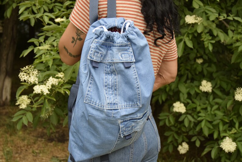 May include: A blue denim backpack with a drawstring closure and multiple pockets. The backpack is made from recycled denim and has a unique, handmade look.