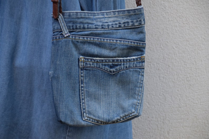 Puede incluir: Un bolso bandolera de mezclilla azul con cierre de solapa y un bolsillo grande en la parte delantera. El bolso tiene una correa de cuero marr&oacute;n.
