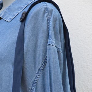 Puede incluir: Una camisa vaquera azul claro con un detalle de correa azul oscuro. La camisa tiene un cuello cl&aacute;sico y una tapeta de botones. Las correas azul oscuro est&aacute;n unidas a la camisa con una hebilla de pl&aacute;stico negro.