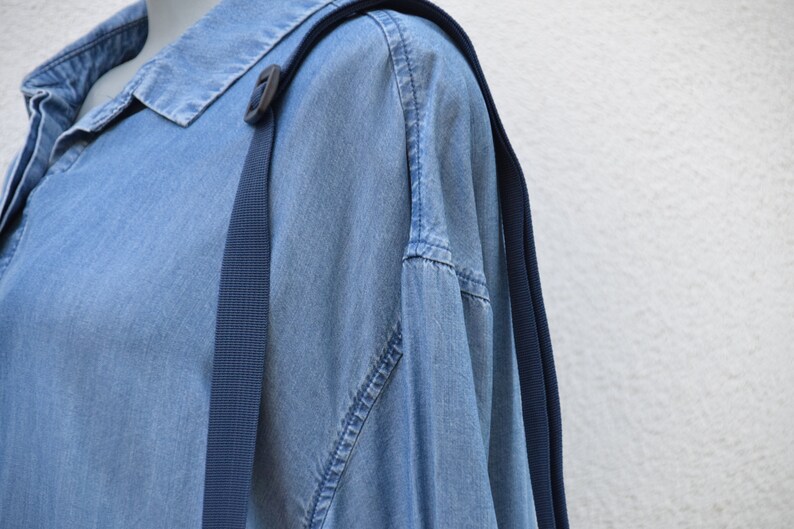 Puede incluir: Una camisa vaquera azul claro con un detalle de correa azul oscuro. La camisa tiene un cuello cl&aacute;sico y una tapeta de botones. Las correas azul oscuro est&aacute;n unidas a la camisa con una hebilla de pl&aacute;stico negro.