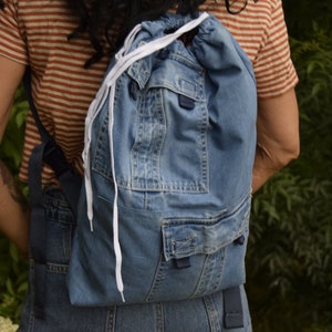 May include: A blue denim backpack with white drawstrings and a large front pocket. The backpack is made from recycled denim.