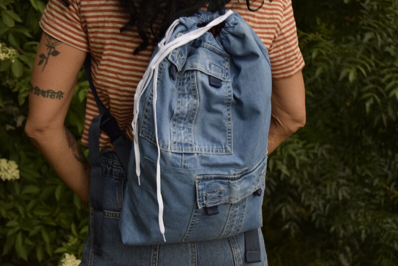 May include: A blue denim backpack with white drawstrings and a large front pocket. The backpack is made from recycled denim.