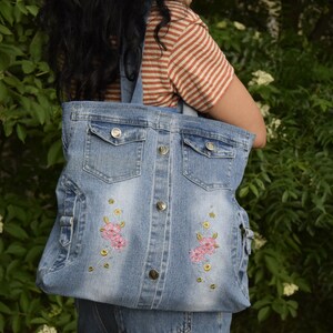 May include: A blue denim tote bag with two front pockets and embroidered pink flowers. The bag has a button closure.