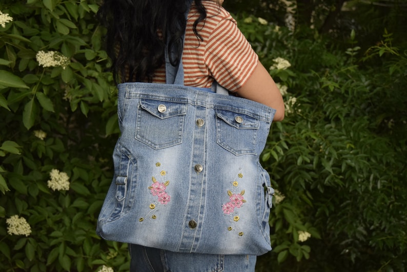 May include: A blue denim tote bag with two front pockets and embroidered pink flowers. The bag has a button closure.