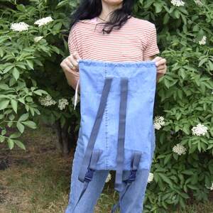 May include: A blue denim backpack with adjustable straps. The backpack is being held by a person wearing a striped shirt.