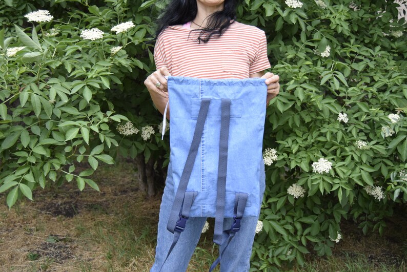May include: A blue denim backpack with adjustable straps. The backpack is being held by a person wearing a striped shirt.