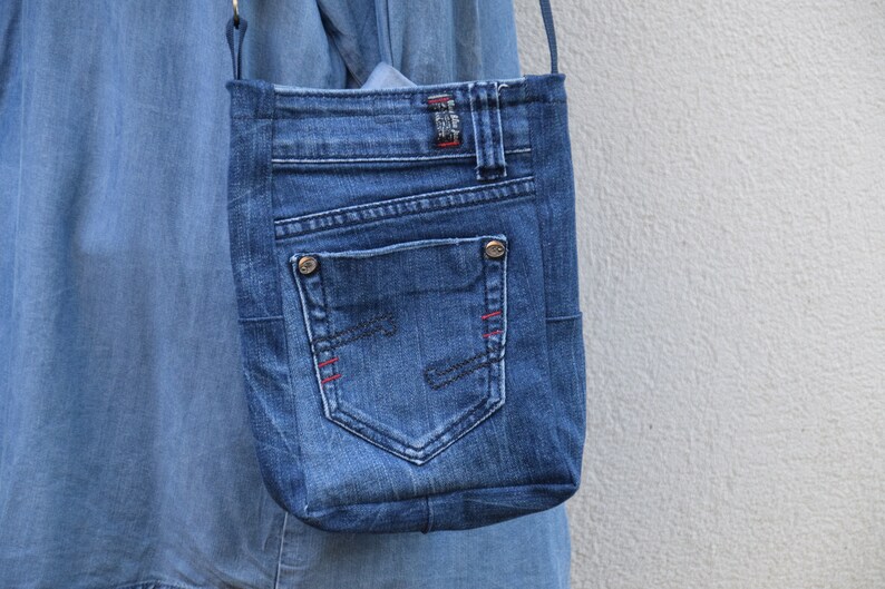 Puede incluir: Bolso cruzado de mezclilla azul con un bolsillo y detalles de costura rojos. El bolso est&aacute; hecho de jeans reciclados y presenta los detalles originales del bolsillo y la trabilla. El bolso se suspende con una correa fina.