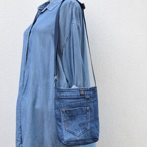 Puede incluir: Un bolso cruzado de mezclilla con una correa larga y delgada. El bolso est&aacute; hecho de jeans azules reciclados, con bolsillos, costuras y un bot&oacute;n de metal. El bolso cuelga contra un vestido camisero de mezclilla azul claro.