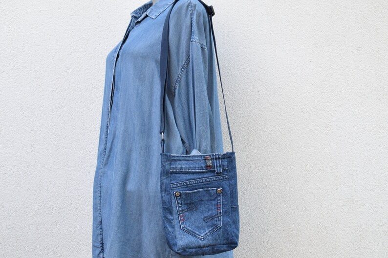 Puede incluir: Un bolso cruzado de mezclilla con una correa larga y delgada. El bolso est&aacute; hecho de jeans azules reciclados, con bolsillos, costuras y un bot&oacute;n de metal. El bolso cuelga contra un vestido camisero de mezclilla azul claro.