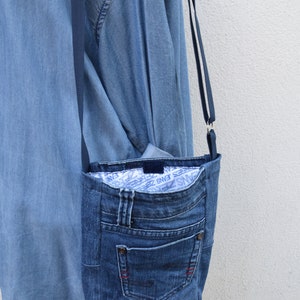 Puede incluir: Un bolso cruzado de mezclilla hecho de jeans reciclados. El bolso tiene un bolsillo frontal, un forro interior azul con texto blanco y una correa larga y delgada. El bolso es de color azul medio y cuelga contra una pared blanca.