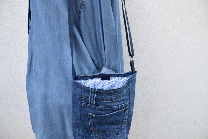 Puede incluir: Un bolso cruzado de mezclilla hecho de jeans reciclados. El bolso tiene un bolsillo frontal, un forro interior azul con texto blanco y una correa larga y delgada. El bolso es de color azul medio y cuelga contra una pared blanca.