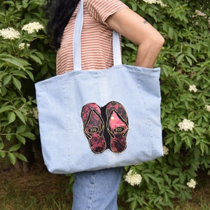 May include: A light blue denim tote bag with a pair of pink and black flip flops embroidered on the front. The flip flops have gold lettering that says "Rio" on each strap.