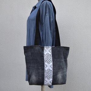 May include: A denim tote bag with a white lace trim down the center. The bag has black straps and is made from recycled denim.