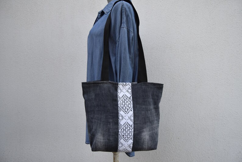 May include: A denim tote bag with a white lace trim down the center. The bag has black straps and is made from recycled denim.